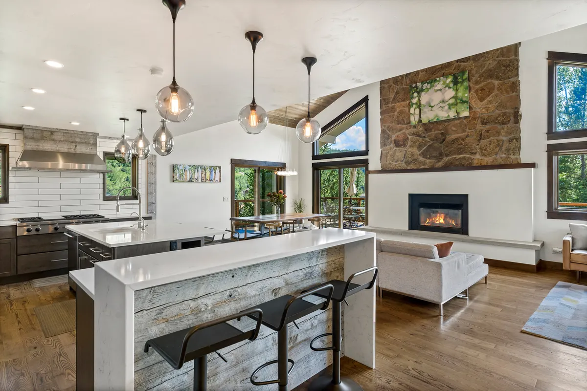 The image features a spacious open-plan interior with a large kitchen island perfect for gathering, a cozy fireplace, expansive windows framing mountain views, and tasteful artwork adorning the walls. The atmosphere is warm, inviting, and ideal for relaxing or entertaining with friends and family.