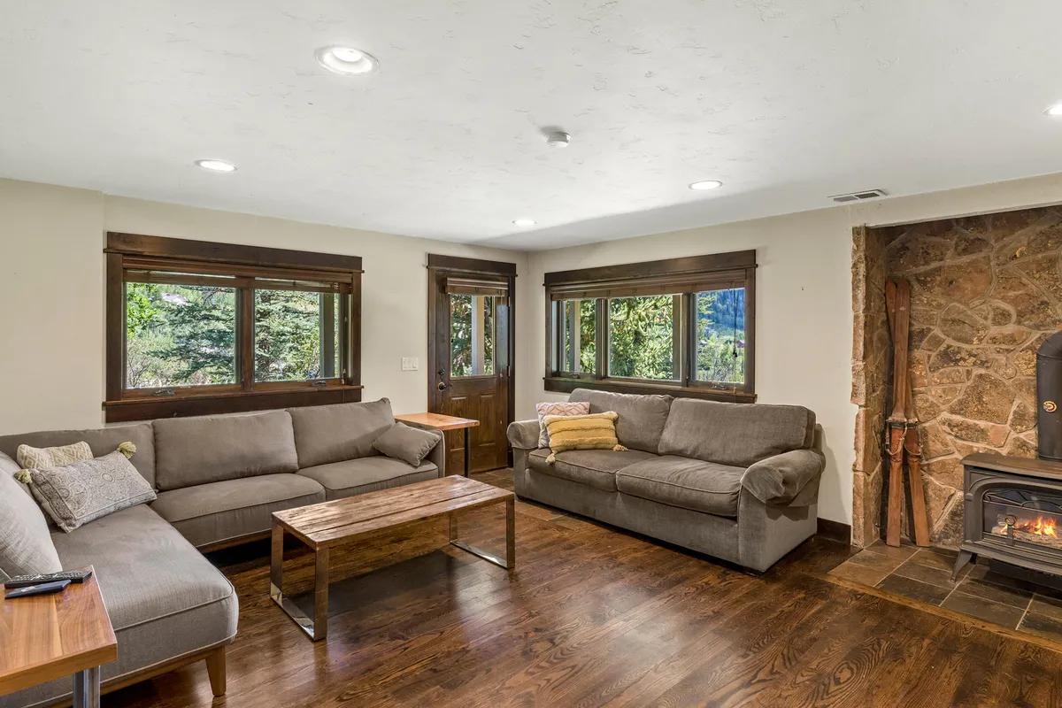 A cozy interior living area with sofas arranged around a fireplace, coffee table, and windows that look out toward the mountain views. The layout encourages relaxed socializing after a day of adventure.