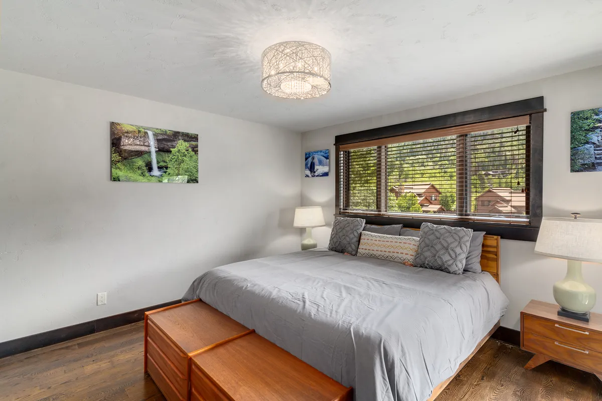 This image captures a serene bedroom with a comfortable bed dressed in gray bedding, large window with wooden blinds allowing in soft natural light, and tasteful artwork on the walls. Nightstands with lamps on either side create an inviting, restful atmosphere.