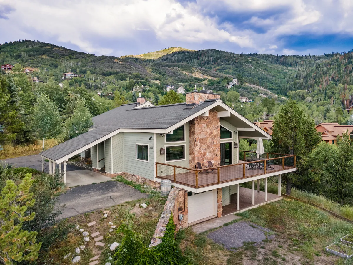 A wider exterior perspective captures the wooden deck with railing, a stone chimney, large windows, and the stunning mountain scenery beyond. This angle expands the context, showing how the deck wraps around the home and connects directly with the natural landscape, suggesting multiple access points including from the main bedroom.