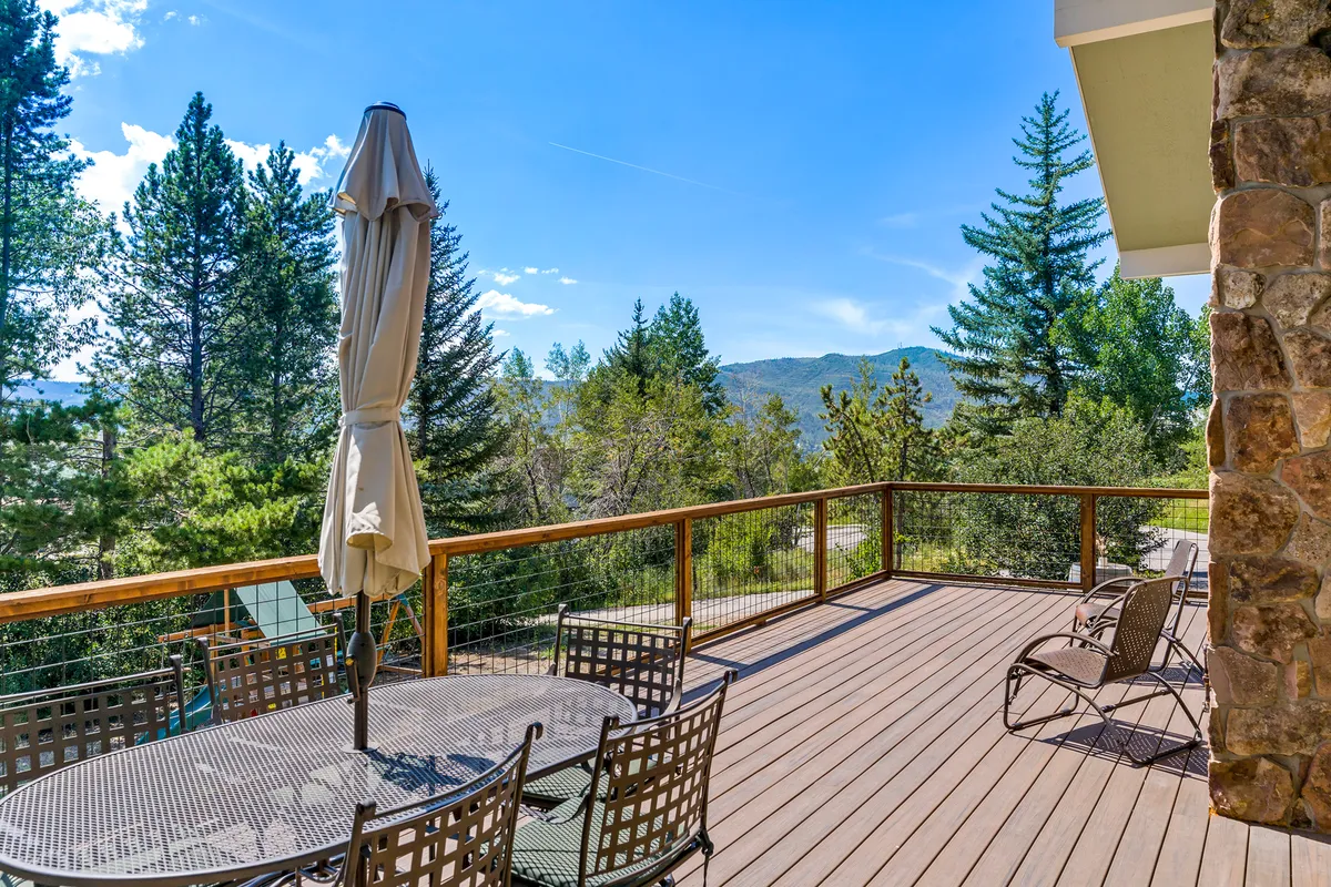 A serene shot of the deck featuring a dining table under an umbrella, surrounded by lush trees and a picturesque view of the mountains. The deck railing and the setup evoke feelings of privacy, tranquility, and celebration under the open sky. The setting is ideal for both intimate and social occasions, in harmony with the mountain lifestyle described.
