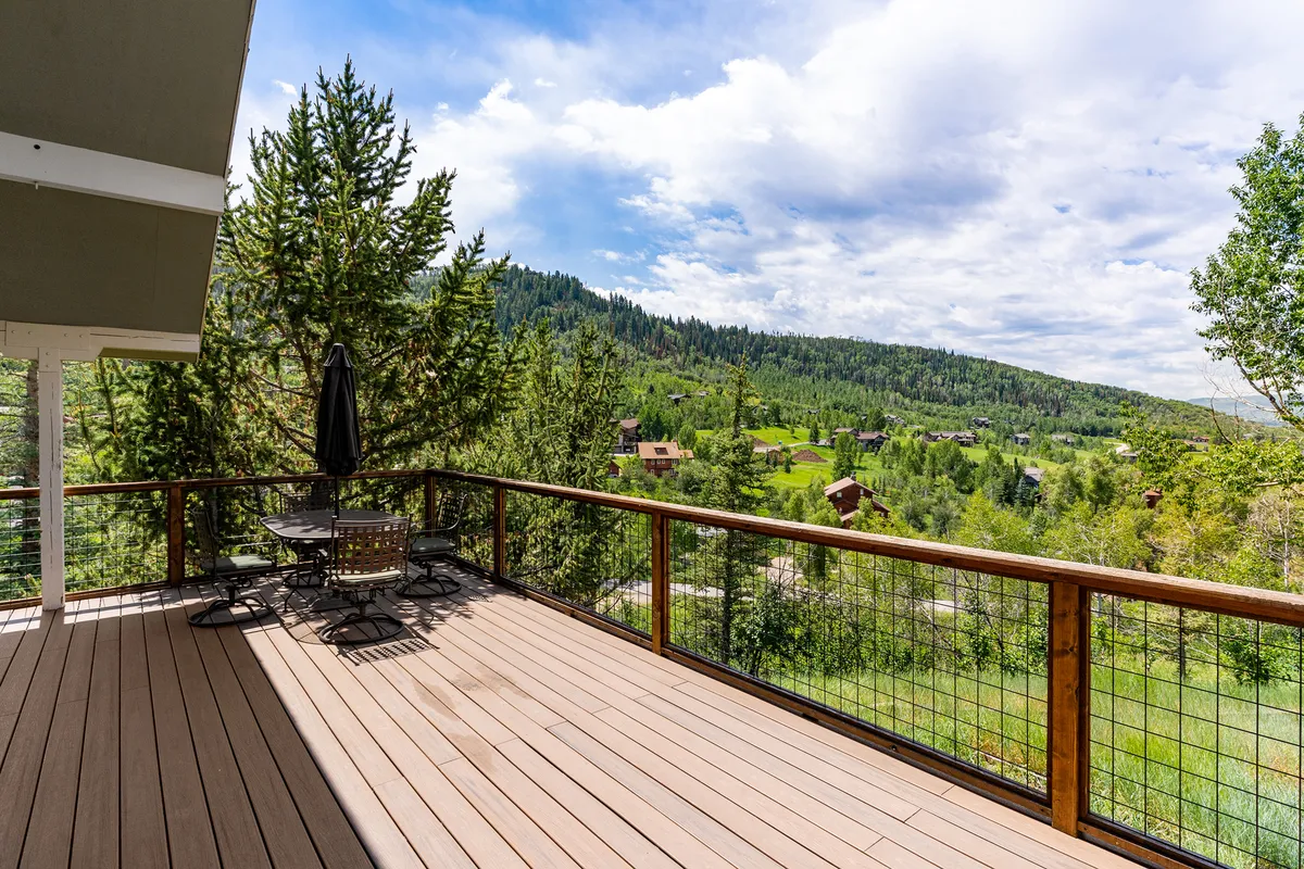 A peaceful deck featuring comfortable outdoor furniture, a railing with a clear sightline to dramatic mountain views, and a backdrop of tall trees. The scene invites moments of relaxation, outdoor dining, or simply admiring the breathtaking landscape at sunrise or dusk.