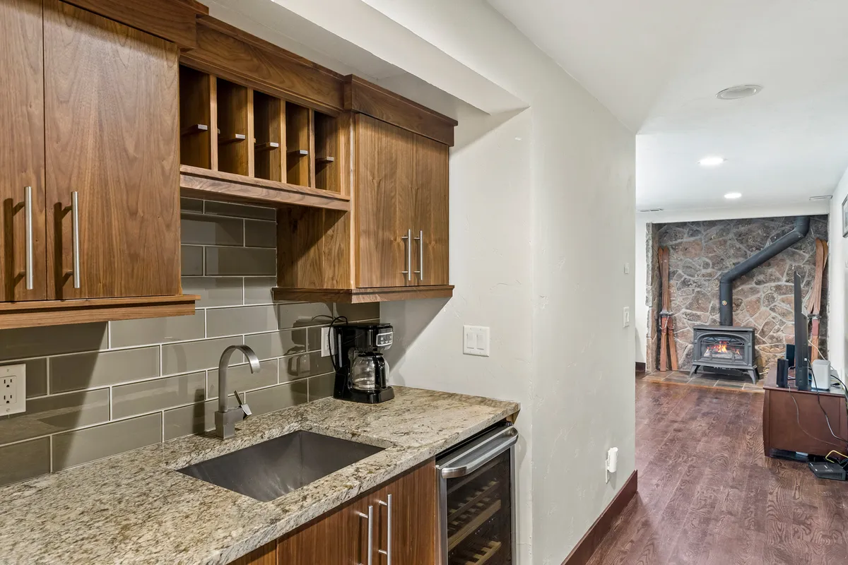 Featuring a granite countertop, wood cabinetry, coffee maker, wine fridge, and a stone fireplace, this modern space blends functionality with cozy charm. It invites guests to enjoy refreshments or casual snacks in a stylish wet bar setting, with the warmth of a nearby fireplace.