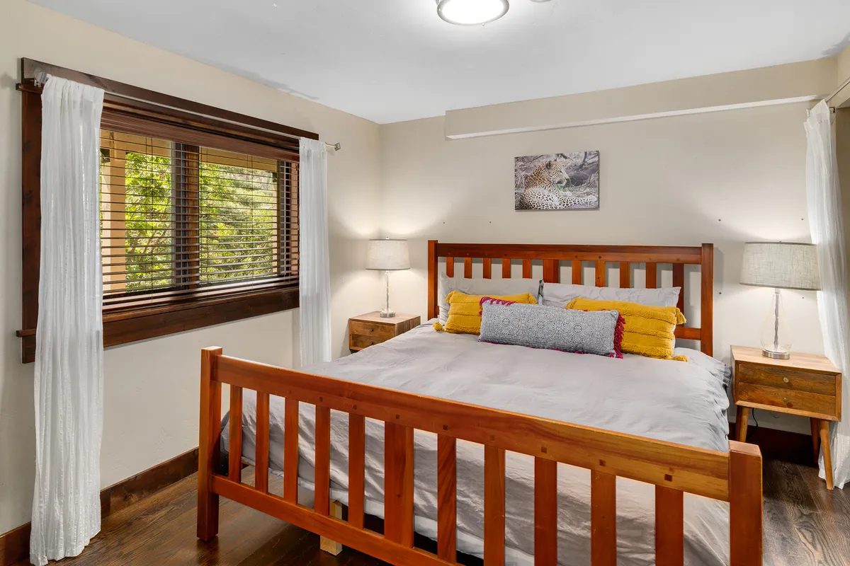 A warmly lit bedroom featuring a sturdy wooden bed frame adorned with decorative pillows, flanked by bedside lamps, with a window allowing natural light to filter in. The space feels inviting and restful—ideal for unwinding after adventurous days.