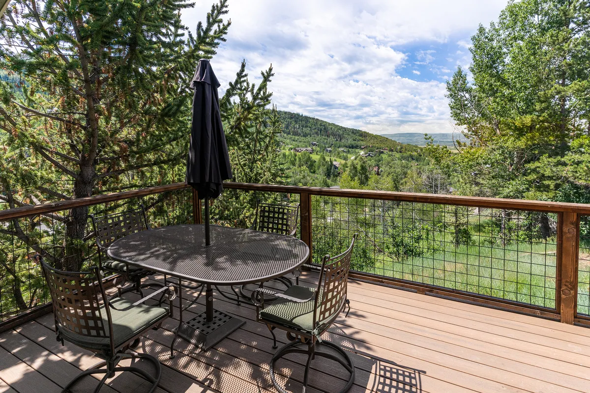 An inviting outdoor dining area with a table and umbrella, surrounded by lush trees and boasting an expansive mountain view. The scene is set for relaxed meals, morning coffee, or evening gatherings as the sun rises or sets behind the peaks.